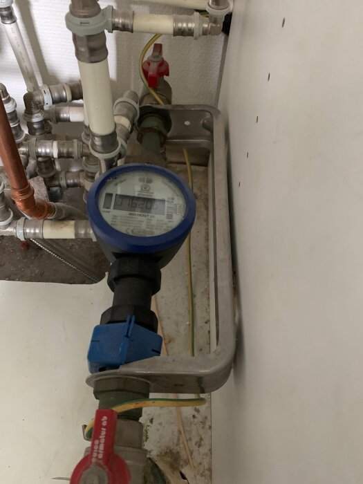 Vattenmätare monterad på en metallkonsol omgivet av flera rör och ventiler i ett trångt utrymme.