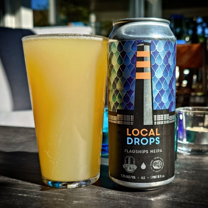 Ett glas med ljus öl bredvid en burk märkt "Local Drops Flagships NEIPA" på ett bord, förmodligen från St Paul, MN.