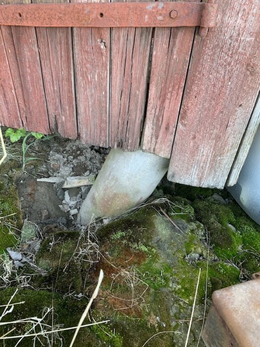 Rostig trägrund med en lutande betongplint, omgiven av mossa och jord.