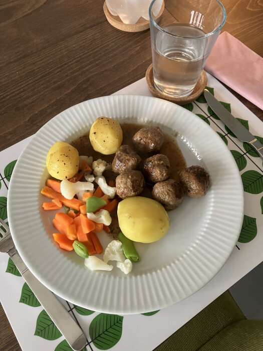 Tallrik med köttbullar, potatis, sås och mixade grönsaker. Glas med vatten bredvid på en bordstablett med bladmotiv.