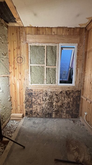 Trärum med isolering och fönster, visar innerväggens panel och reglar för kabeldragning, möjligt för renovering och väggrensning.