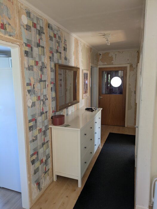 Hallen under renovering med avskalade väggar och synliga gamla tapetrester. En byrå står mot väggen, och en dörr syns i slutet av gången.