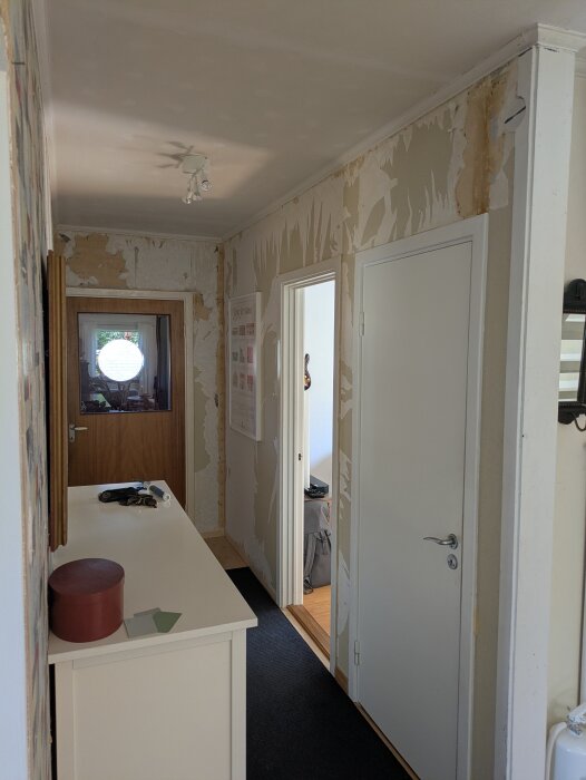 En avlång hall under renovering med delvis borttagen tapet, synliga fläckar och nedsatt gipskartong runt flera dörröppningar.