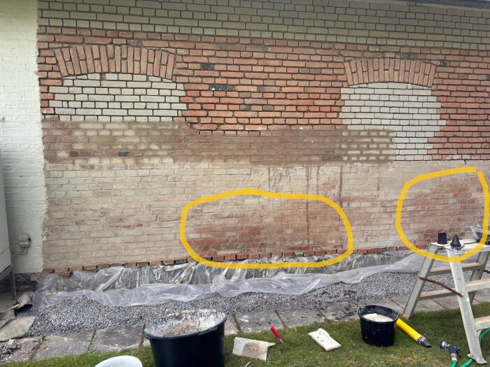 Tegelvägg med fuktfläckar inringade i gult; plastskydd och verktyg vid de dränerade grusfyllda områdena framför väggen.