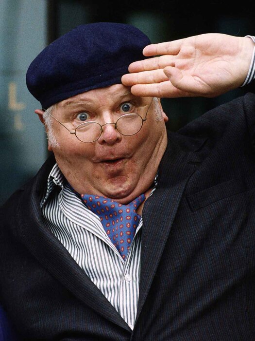 Man med glasögon, svart basker, randig skjorta och blå slips, gör en uttrycksfull pose och håller handen vid ögonbrynen.