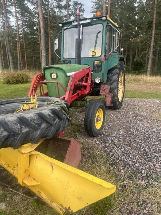 Äldre John Deere-traktor med gul plog i förgrunden, omgiven av tallskog och grusväg.