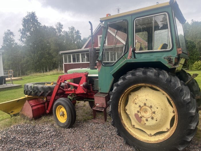 Äldre John Deere-traktor, grön med gul fälg, står på en grusväg framför ett rött hus, omgiven av träd och gräs.