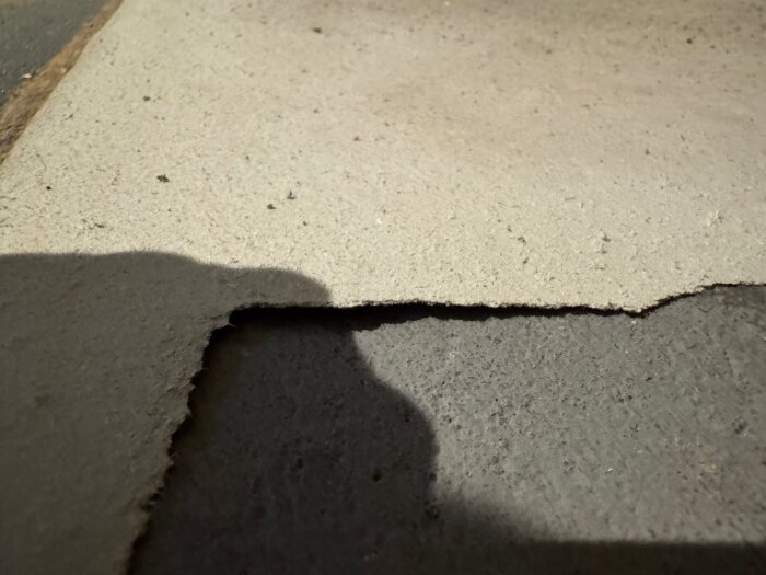 Vit pappliknande yta under plastmatta på betonggolv, potentiellt asbestpapp från hus byggt 1965, ses mot bakgrund av skuggor och ojämn yta.