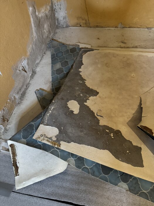 Uppbruten plastmatta som visar lager av vit papp/filt ovanpå blåmönstrat golv i källarförråd, potentiellt asbestinnehåll.
