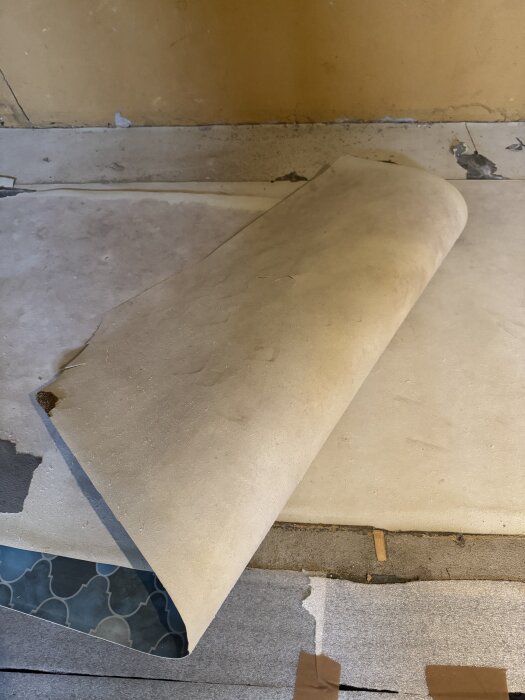 Riven plastmatta uppvikt över vit pappliknande massa, potentiellt asbestinnehållande, på betonggolv i förråd från 1965.