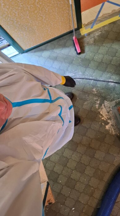 Person i skyddskläder arbetar på ett golv med grön mönstrad matta, omgiven av verktyg och tejpade plastskydd, vid renovering med asbestrisk.