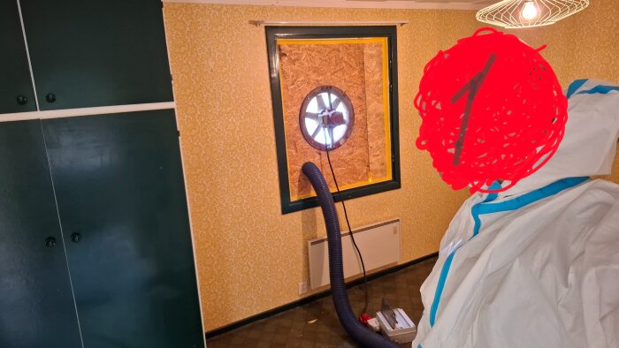 Rum med gul tapet, svart fläkt monterad i fönstret för ventilation, vitt skyddsdräkt, svart skåp och arbetsutrustning på golvet.