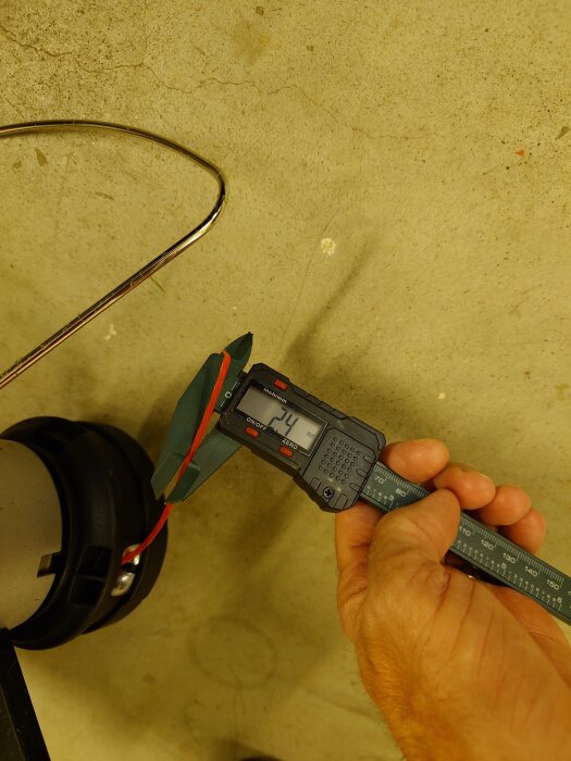 Hand håller en digital skjutmått som mäter en röd kabel till 2,4 mm på ett beige underlag.
