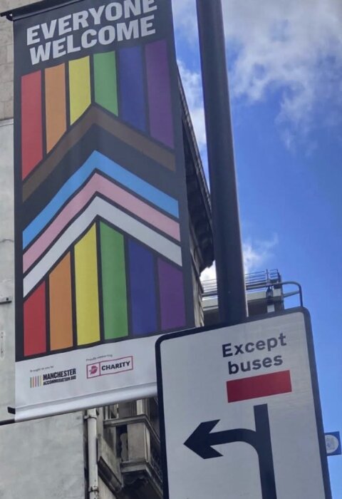 Skylt med "Everyone Welcome" och regnbågsfärger på en lyktstolpe, bredvid en trafikskylt med texten "Except buses" och en vänstersvängpil.