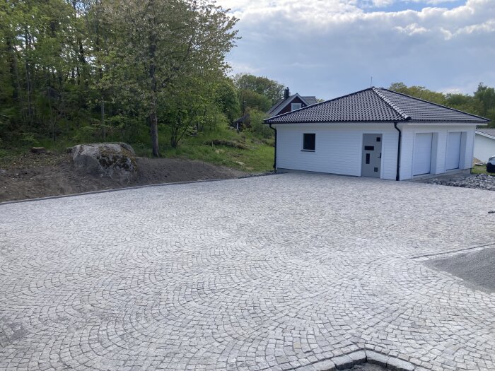 En nyanlagd stenlagd uppfart framför ett modernt vitt garage med dubbeldörrar, omgiven av grönska och träd i bakgrunden.