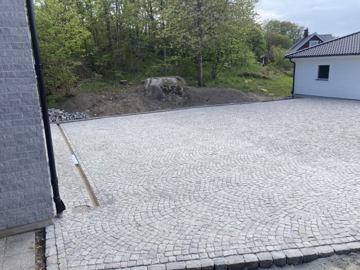 En nyanlagd stenbelagd uppfart framför ett hus med omgivande grönska och en stor sten i bakgrunden.