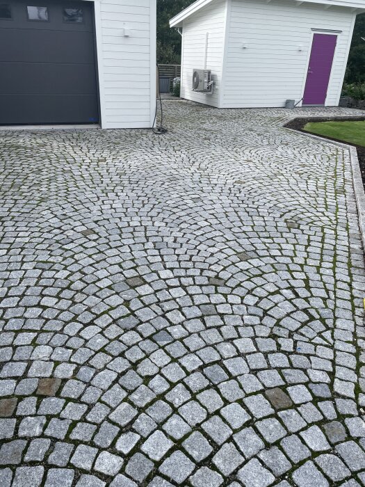 Uppfart med mönstrad stensättning i grå nyanser framför garage och förråd, med grönska mellan stenarna.