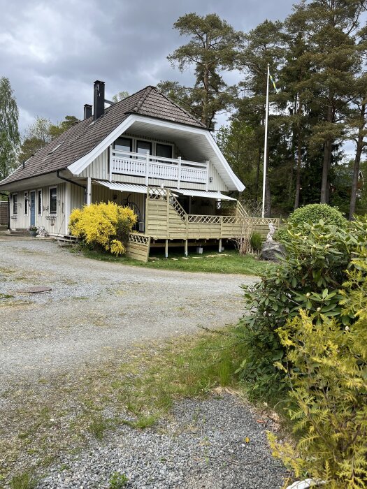 Vit villa med träbalkong, omgiven av gräsmatta, buskar och en grusad uppfart. En svensk flagga syns vid flaggstången, en mulen dag.