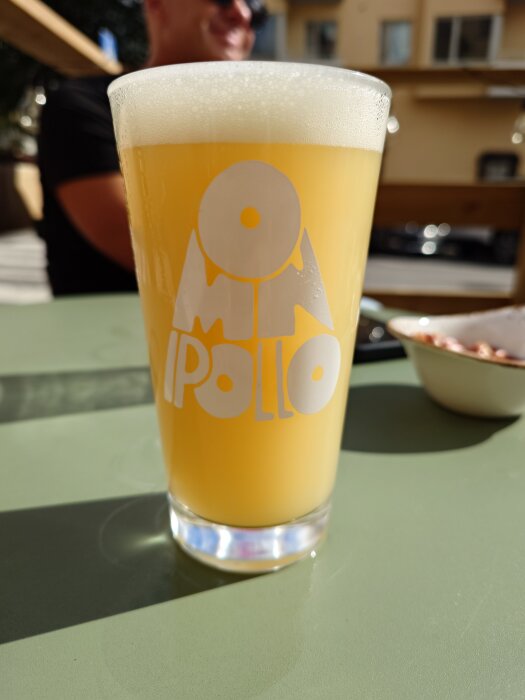 Ett glas fyllt med en ljus, skummig IPA står på ett bord i solljus; Omnipollo-logga på glaset.