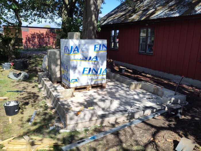 Byggarbetsplats med pall märkta "Finja" placerad på betonggrund intill en röd byggnad, omgiven av byggmaterial och verktyg.