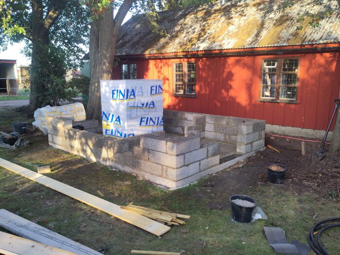 Byggprojekt med blockgrunder utanför rött hus, material från Finja, omgivna av träd och verktyg på marken.
