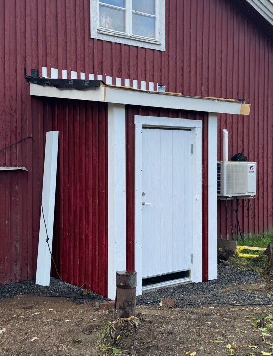 Smått rött pumphus med vitt dörrfoder, byggt mot en röd husgavel. En luftkonditioneringsenhet är monterad på väggen.