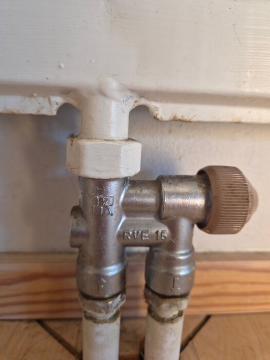 Radiatorventil med två rör och brunt ställdon i källarplan, märkt "RVE 15", monterad mot en vit vägg och trälist.