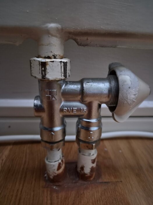 Radiatorventil i källarplan med vita ställdon installerad på träbeklädd golv, med synliga värmerör och viss slitage.