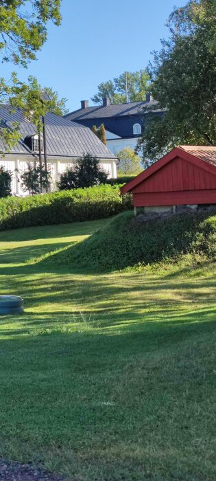 En röd träbod i en grönskande trädgård med klippt gräs och en större vit byggnad i bakgrunden, under en klarblå himmel.