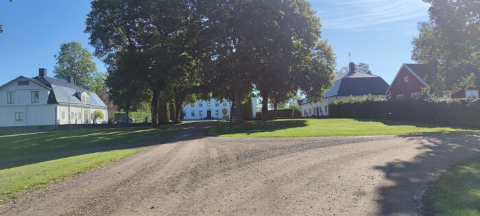 Vägkorsning framför tre hus, omgiven av stora träd och välklippt gräsmatta under en klarblå himmel.