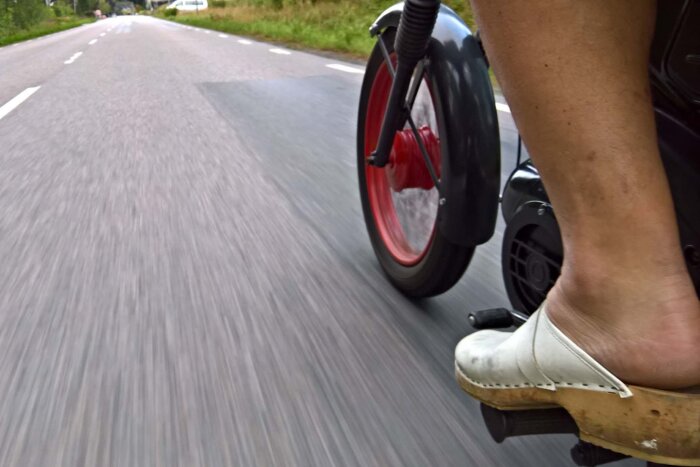 Fot på mopedpedal iförd vit träsko, röd hjulnav syns, moppen körs på landsväg.