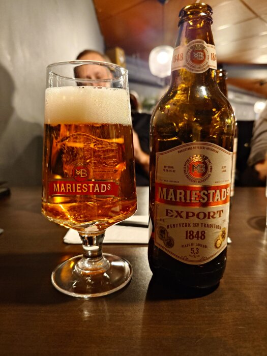 Glas och flaska med Mariestads öl på ett bord inomhus.