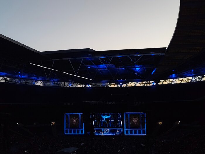 Konsert på Wembley Stadium med scen och blå belysning under kvällshimmel.