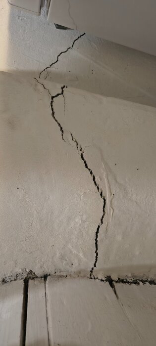 Spricka i vitmålad vägg vid golvkant, troligen orsakad av sättningar i huset.