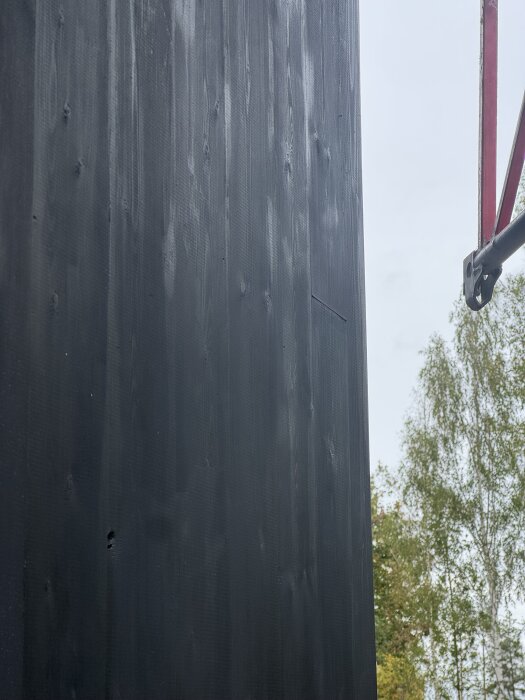 Svartmålad träpanel med byggställning utomhus och träd i bakgrunden.