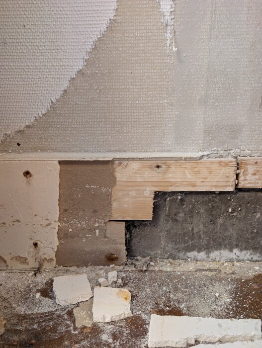 Skadad garagevägg med gips och papp som faller sönder, synliga trä- och betongdelar, samt ett hål i träet och gipsskivor på golvet.