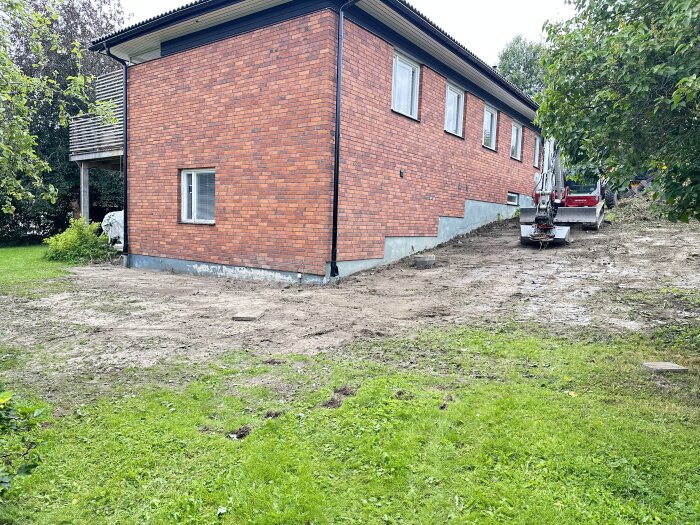 Tegelhus med grävmaskin bredvid på en lerig mark; potentiell byggarbetsplats under utveckling.