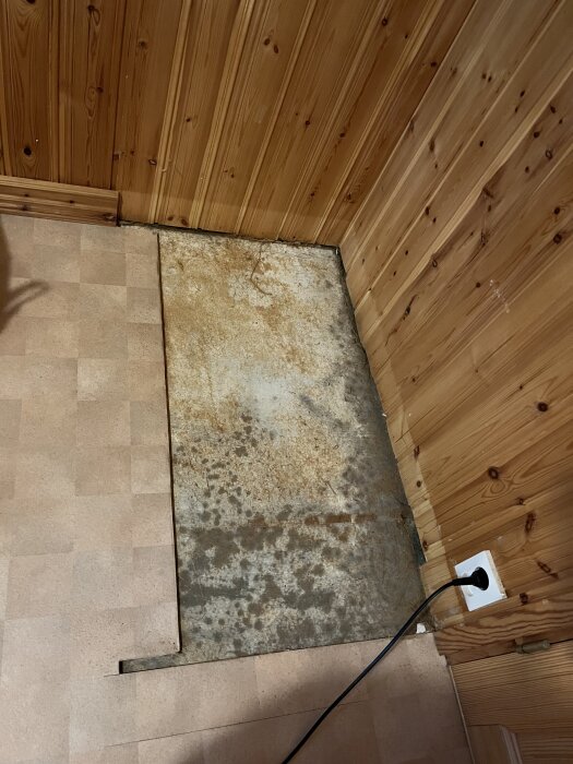 Golv med borttagen plastmatta visar mörka fläckar och spånskiva i ett hörn av ett rum med träpanel, intill ett eluttag och kabel.
