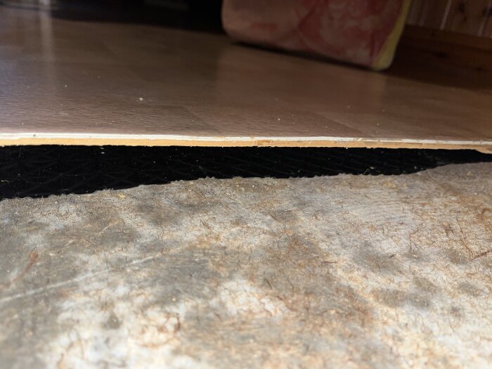 Närbild av golvyta där plastmatta ligger ovanpå spånskiva, med svart plast under, och fläckar på betongplattan syns tydligt.