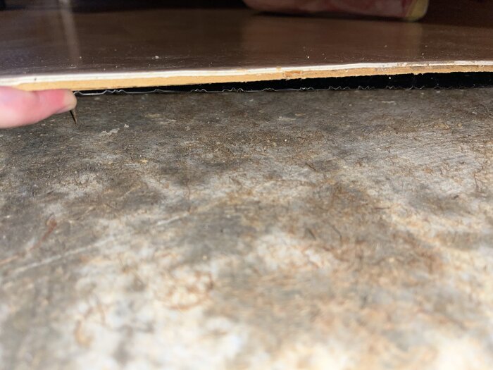 Närbild av ett öppnat golv där en hand lyfter spånskivan, synliggör svart plast och en ojämn yta med rester av juteväv på betongplattan.