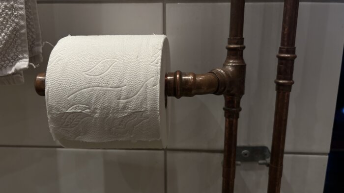 Toalettpappershållare i obehandlat rör i badrum, med papper och handduk synliga.