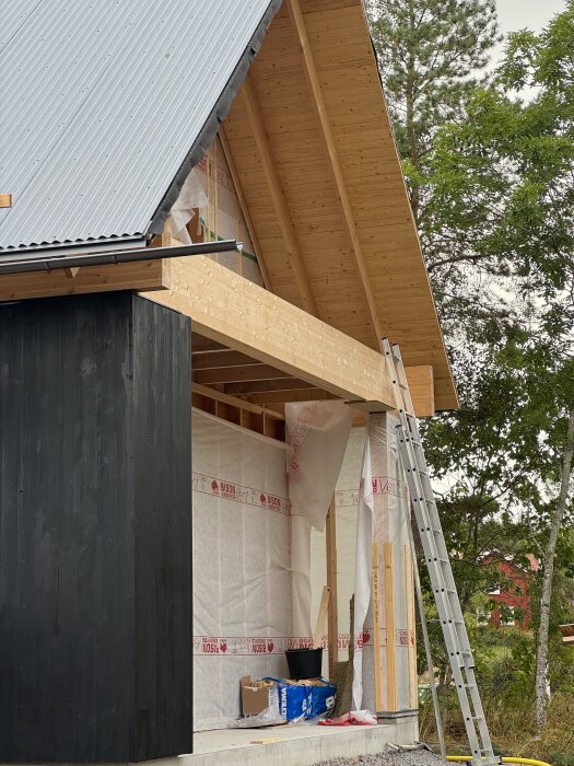 Hus under byggnation med synligt hörn, svart fasadpanel och takstolar i trä. Stege står lutad mot väggen, omgiven av träd.