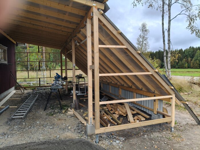 Byggprojekt med pågående takbygge och träkonstruktion utomhus, omgiven av natur och verktyg som stege och såg.