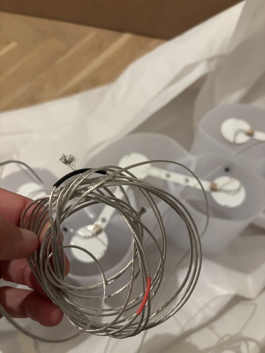En hand håller i en slinga av metalltråd framför vita plastkomponenter på ett träbord. Tråden är delvis isolerad med svart och röd plast.