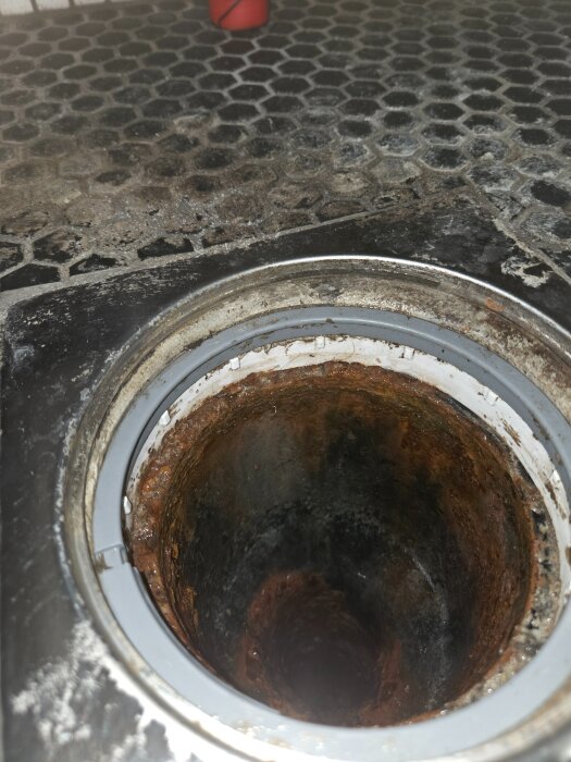 Närbild av ett slitet vattenlås i ett golvavlopp, med rost och en möjlig trasig gummipackning som kan orsaka läckage av avloppslukt.