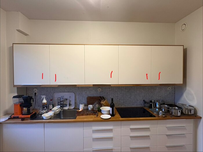 Kök med vita skåpluckor, märkta med röda markeringar där nya handtag planeras. Diskbänk med glas och köksredskap framför kaklad vägg.