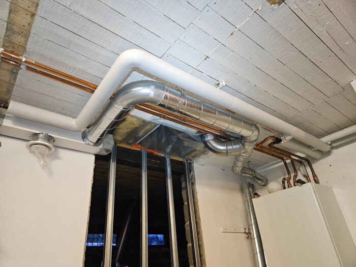 Ventilationsrör i metall och plast monterade i taket på ett byggprojekt, med flera böjar och anslutningar till ett vitt skåp på väggen.