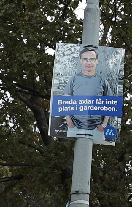 Valskylt med texten "Breda axlar får inte plats i garderoben" fäst på en lyktstolpe.