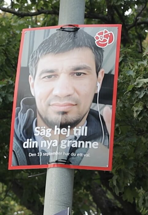 Affisch från Socialdemokraterna med texten "Säg hej till din nya granne" och datum för val, fäst på en stolpe med träd i bakgrunden.