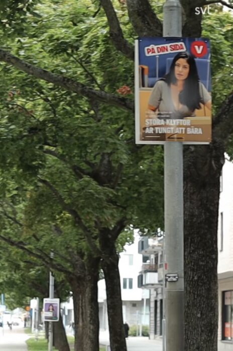 Valaffisch för Vänsterpartiet på lyktstolpe längs trädallé med texten "Stora klyftor är tungt att bära", bakom syns byggnader och trottoar.